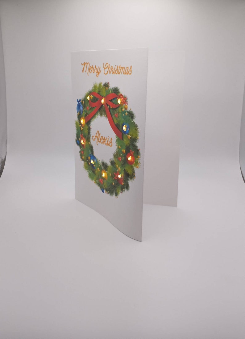 K&ouml;nnte beinhalten: Wei&szlig;e Weihnachtskarte mit einem festlichen Kranzdesign. Der Kranz ist mit einer roten Schleife, bunten Lichtern und blauen Ornamenten verziert. Auf der Karte stehen die Worte "Merry Christmas" und der Name "Alexis".