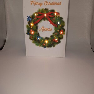 K&ouml;nnte beinhalten: Eine wei&szlig;e Weihnachtskarte mit den Worten "Merry Christmas" in orangefarbener Schrift. Ein gr&uuml;ner Kranz mit roter Schleife und Ornamenten umgibt das Wort "Alexis". Der Kranz hat kleine Lichter.