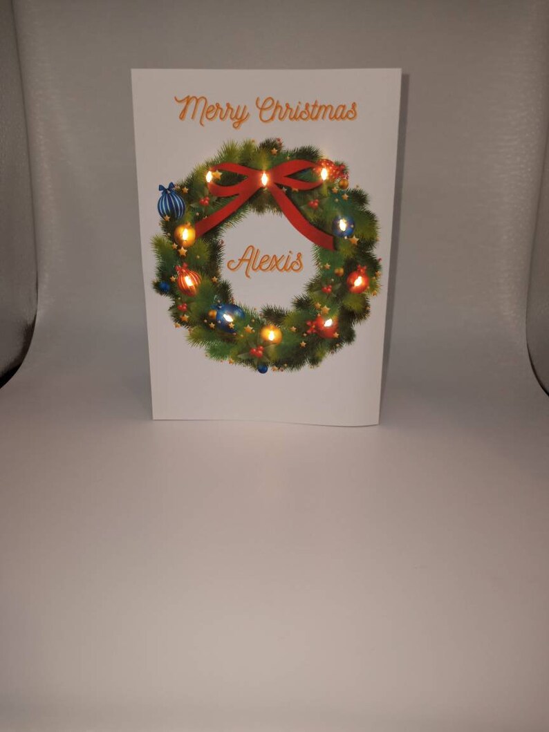 K&ouml;nnte beinhalten: Eine wei&szlig;e Weihnachtskarte mit den Worten "Merry Christmas" in orangefarbener Schrift. Ein gr&uuml;ner Kranz mit roter Schleife und Ornamenten umgibt das Wort "Alexis". Der Kranz hat kleine Lichter.