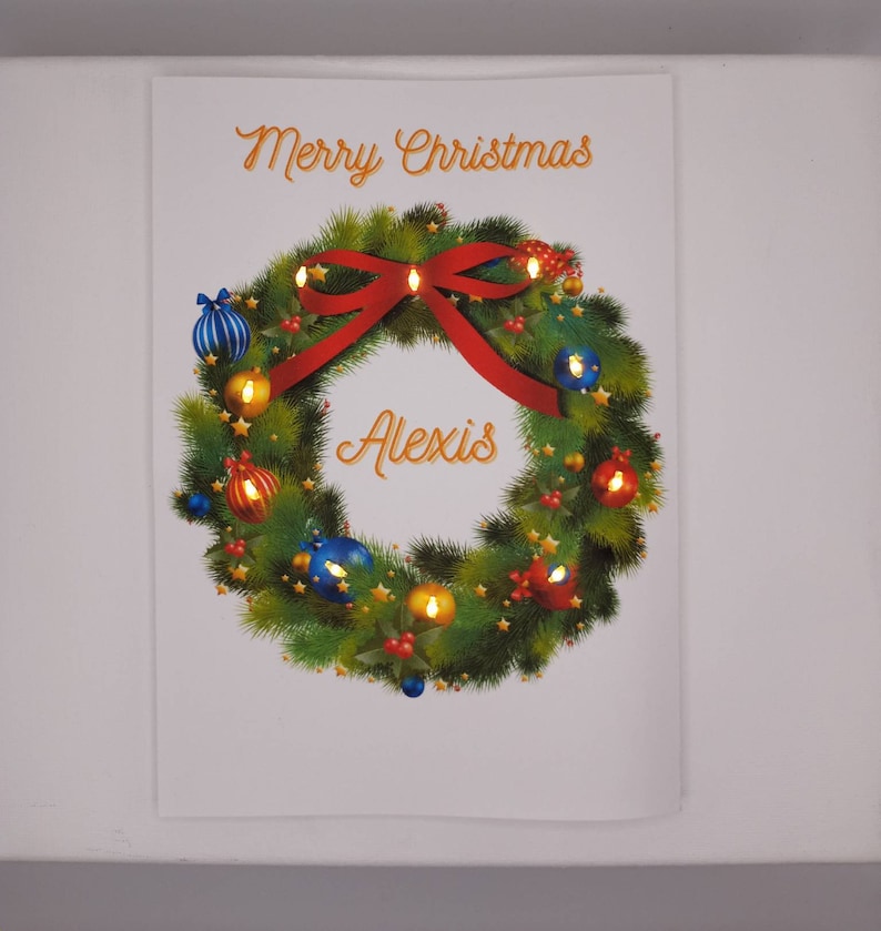 K&ouml;nnte beinhalten: Weihnachtliches Bild mit einem festlichen Kranz, einem roten Bogen und Ornamenten. &Uuml;ber dem Kranz steht &bdquo;Merry Christmas&ldquo;, und der Name &bdquo;Alexis&ldquo; befindet sich in der Mitte.