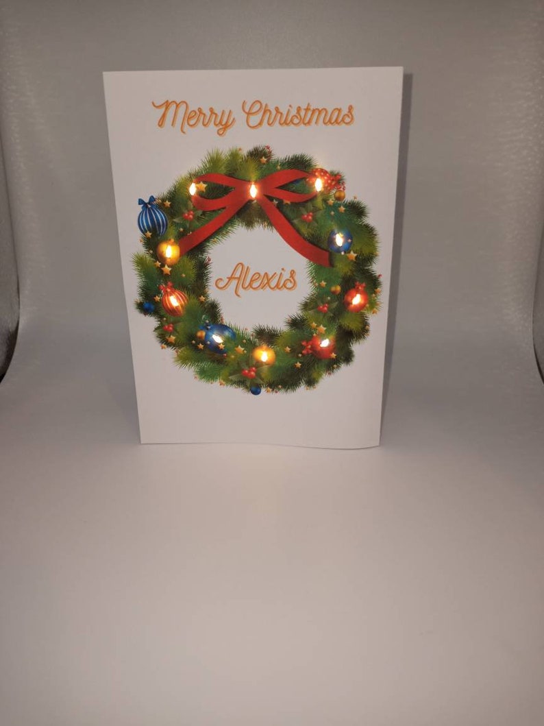 K&ouml;nnte beinhalten: Eine wei&szlig;e Weihnachtskarte mit einem festlichen Kranz, einer roten Schleife und bunten Lichtern. Auf der Karte stehen die Worte "Merry Christmas" und der Name "Alexis". Der Kranz ist mit blauen und goldenen Kugeln geschm&uuml;ckt.