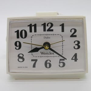 May include: A rectangular white Westclox electric alarm clock with a clear plastic face cover. The clock features a white dial with large black Arabic numerals from 1 to 12, black hour and minute hands, a gold second hand, and the text Dialite.