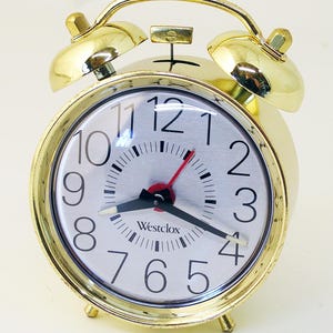 May include: A gold-colored Westclox alarm clock with a round face and two bells on top. The clock face is silver with black numbers and hands, and a red second hand. The clock has a classic design and is set on a white surface.