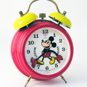 May include: Red alarm clock with yellow bells featuring a Mickey Mouse illustration on the face. The clock face reads "Mickey Mouse" and "Walt Disney Productions" with a Mickey Mouse image in the center.
