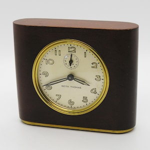 May include: A rectangular Seth Thomas desk clock with a dark brown wooden casing and a gold-toned metal bezel. The clock face is off-white with black Arabic numerals, black hands, and a small sub-dial at the top. The text MADE IN USA is visible.