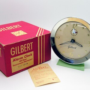 May include: A vintage Gilbert alarm clock with a silver-tone bezel and a tan face. The clock has black hands and gold-colored numerals. The clock sits on a green base. A red box with the Gilbert logo and text is next to the clock.