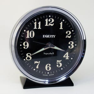 May include: A silver alarm clock with a black face and white numbers. The clock says "EQUITY Superbell" and "HONG KONG".