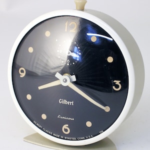 May include: White alarm clock with a black face and gold numerals. The clock is labeled "Gilbert Luminous" and "GILBERT CLOCKS MADE IN WINSTED, CONN. U.S.A. 176".