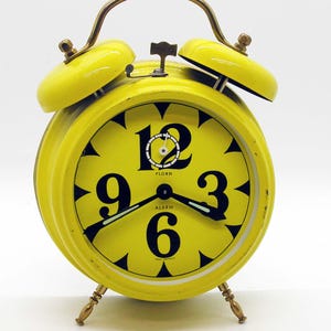 May include: A vintage yellow alarm clock with a round face and a metal handle. The clock face features large black numerals and hands, with the word "ALARM" printed below the number 6. The clock has two bells on top and stands on four small legs.