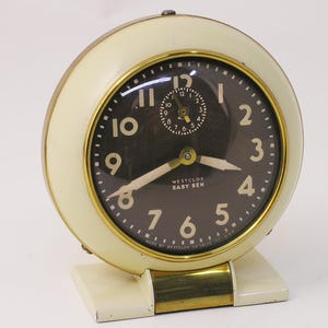 May include: Cream-colored vintage "Baby Ben" alarm clock with a black face and gold accents. The clock features large white numerals, a smaller inner dial, and the text "WESTCLOX BABY BEN". The clock is approximately 4 inches tall.
