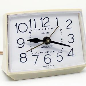 May include: A vintage Westclox dialite alarm clock with a cream-colored plastic case. The clock face is white with black numerals and hands. The clock reads "dialite" and "WESTCLOX" and is marked "MADE IN U.S.A."