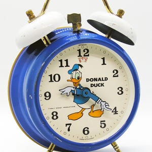 May include: Vintage blue and white alarm clock featuring Donald Duck. The clock has a gold handle and legs, with a white face and black numbers. The clock face reads "DONALD DUCK" and "WALT DISNEY PRODUCTIONS."