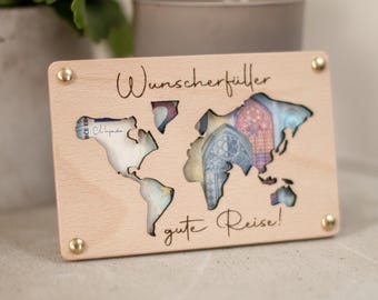 Könnte beinhalten: Eine rechteckige Holzplatte in hellem Farbton mit einer ausgeschnittenen Weltkarte. Der Schriftzug "Wunscherfüller" ist über der Karte eingraviert, "gute Reise!" darunter. Vier goldfarbene Metallschrauben befestigen die Ecken der Platte.