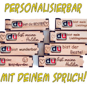 Könnte beinhalten: Ein Stapel aus sechs Holzkisten mit einem Herz und dem Wort "du" auf jeder Kiste. Die Kisten sind mit verschiedenen Botschaften personalisiert, wie z. B. "du bist die BESTE!" und "du bist mein Lieblingsmensch!!". Die Kisten sind übereinander gestapelt und der Text "MIT DEINEM SPRUCH!" steht unter den Kisten.