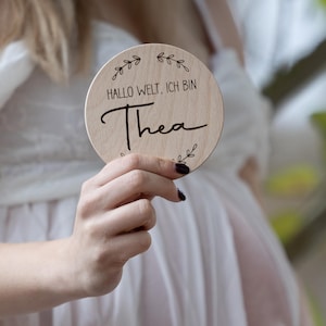 Könnte beinhalten: Ein rundes Schild aus Holz mit dem Text "Hallo Welt, ich bin Thea" in schwarzer Schrift. Das Schild ist mit kleinen Blattmotiven verziert.