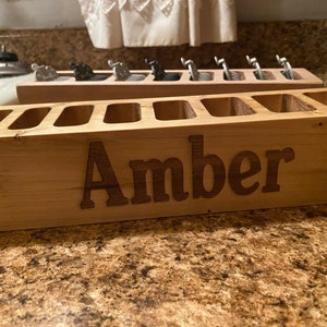 May include: Two wooden organizers, one with the name "Amber" carved into the front. Each has multiple slots for holding items. The top organizer holds silver and black objects. The wood has a natural finish.