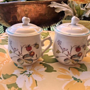 May include: Two white ceramic mugs with lids, featuring a strawberry and floral design. Each mug has a handle and a heart-shaped knob on the lid. The mugs are on a floral patterned surface.