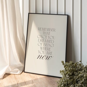 Peut inclure: Impression encadrée en noir avec le texte "Remember that once you dreamed of being where you are now" sur fond blanc. L'impression est appuyée contre un mur blanc avec un rideau blanc en arrière-plan.