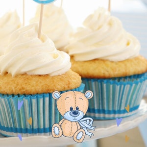 May include: Blue and white cupcakes with white frosting decorated with blue and white baby shower toppers. The toppers say "Baby" and "It's a Boy!" A teddy bear is in the foreground.