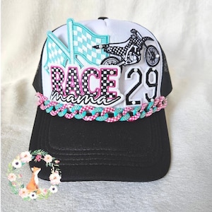 Puede incluir: Una gorra tipo trucker negra y blanca con un parche bordado que muestra una bandera a cuadros, una motocicleta y el texto RACE mama 29. El panel frontal está decorado con una cadena de eslabones alternos en color rosa y turquesa.