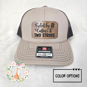May include: Tan and black trucker hat with the text "Fueled by Coffee & Two Stroke" on a brown patch. The hat has a curved brim and an adjustable strap. The hat is a Richardson Original 112 Trucker.
