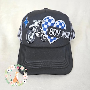 Op de afbeelding: Een zwarte truckerpet met een blauw-wit geblokte hartvormige patch met de tekst BOY MOM, een witte geborduurde afbeelding van een crossmotor en een kleine blauwe vierpuntige ster. De pet heeft een zwarte klep en een zwart-wit geblokte mesh achterkant.