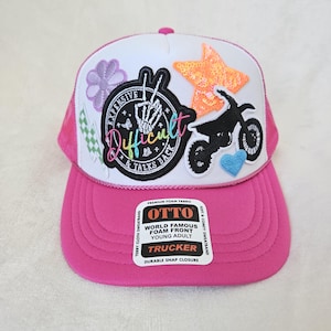 Op de afbeelding: Roze en witte truckerpet met een gebogen klep. De pet heeft geborduurde patches van een motor, ster, bloem en een ronde patch met de woorden "Expensive Difficult & Talks Back". Het heeft een druksluiting.