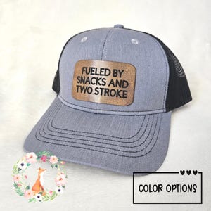 May include: A gray trucker hat with a black mesh back and a curved brim. The hat features a brown leather patch with the text "FUELED BY SNACKS AND TWO STROKE". The hat has a snap closure and is a color option.