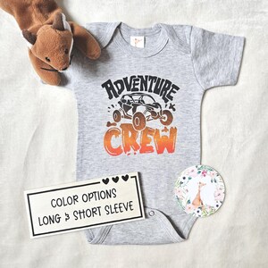 Könnte beinhalten: Ein grauer Baby-Body mit den Worten "ADVENTURE CREW" in Schwarz und Orange, mit einer Grafik eines Geländewagens. Ein kleines Stofftier in Form eines Fuchses und ein floraler Aufkleber liegen neben dem Body. Ein Schild zeigt "COLOR OPTIONS LONG & SHORT SLEEVE."