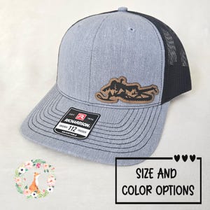 May include: A gray Richardson 112 trucker hat with a black mesh back and a brown leather patch featuring a mountain and snowmobile design. The hat has a curved brim and the text "SIZE AND COLOR OPTIONS".
