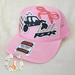 Puede incluir: Una gorra trucker rosa claro con un vehículo todoterreno negro bordado, un corazón a cuadros beige y blanco, un lazo rosa y el texto "hell RZR". Una pegatina en la visera dice "OTTO WORLD FAMOUS FOAM FRONT YOUNG ADULT TRUCKER".