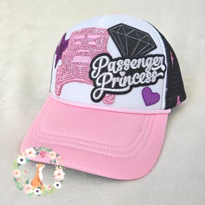 Könnte beinhalten: Eine Trucker-Cap mit rosafarbenem Schirm und weißer Vorderseite, verziert mit gestickten Aufnähern. Das Design zeigt ein glitzerndes rosa Auto, einen schwarzen Diamanten, ein violettes Herz und den Schriftzug Passenger Princess in Schwarz-Weiß.