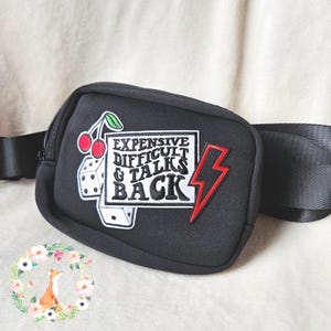 Puede incluir: Riñonera negra con parches bordados que incluyen cerezas, dados y un rayo. El texto en el parche dice "EXPENSIVE DIFFICULT & TALKS BACK". La bolsa tiene una correa negra ajustable.