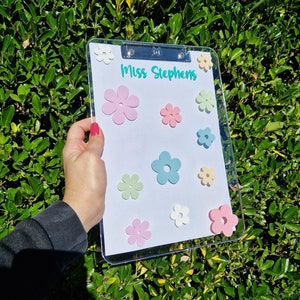 May include: A clear clipboard with a white background and the text "Miss Stephens" in green. The clipboard is decorated with colourful paper flowers in various shades of pink, blue, and yellow.