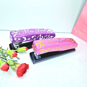 May include: Two personalised staplers with glitter finishes. One is purple with white lettering that says "Mrs. Ford". The other is pink with gold lettering that says "Miss Evelina".