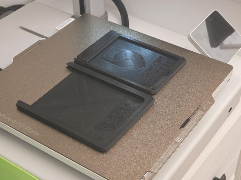 May include: Two black rectangular 3D-printed trays with text and a logo are displayed on a textured, metallic surface. One tray is open, and the other is closed. The text on the trays includes "KPSHIP.COM" and "1088194".
