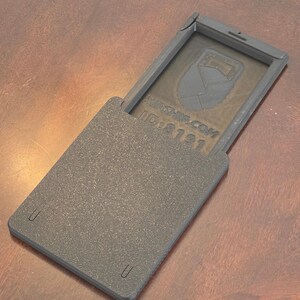 May include: A dark gray, rectangular case with a hinged lid, open to reveal a design and text. The case has a textured surface and rounded corners. The text "ID:8121" is visible. The case is on a brown surface.