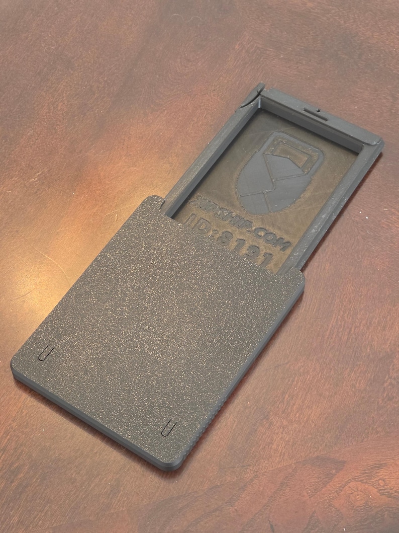 May include: A dark gray, rectangular case with a hinged lid, open to reveal a design and text. The case has a textured surface and rounded corners. The text "ID:8121" is visible. The case is on a brown surface.