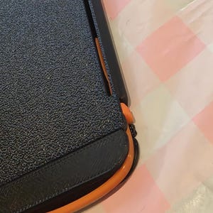 May include: A black and orange paper trimmer with a textured, dark blue surface. The trimmer has a black handle and a bright orange curved edge. The background is a pink and white checkered pattern.