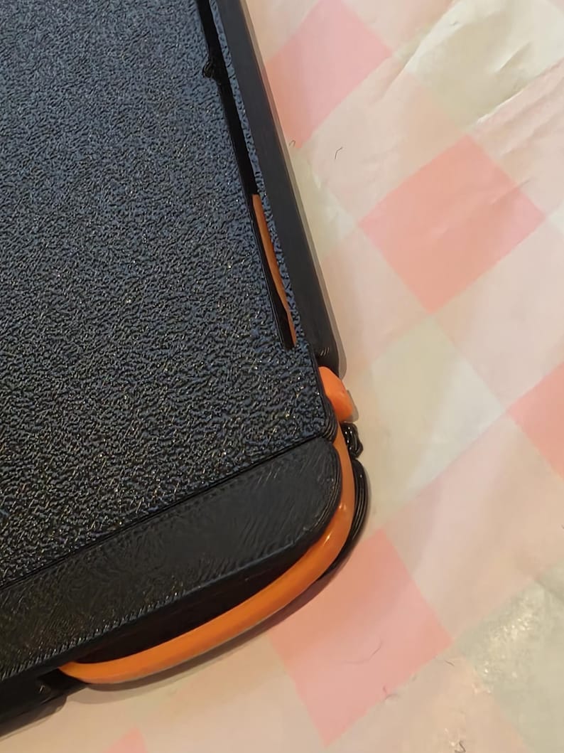 May include: A black and orange paper trimmer with a textured, dark blue surface. The trimmer has a black handle and a bright orange curved edge. The background is a pink and white checkered pattern.