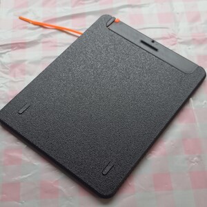 May include: A black, rectangular tablet with a textured surface. The tablet has a small orange cord attached to the top edge. There are two small, recessed slots on the bottom edge and one on the top edge.