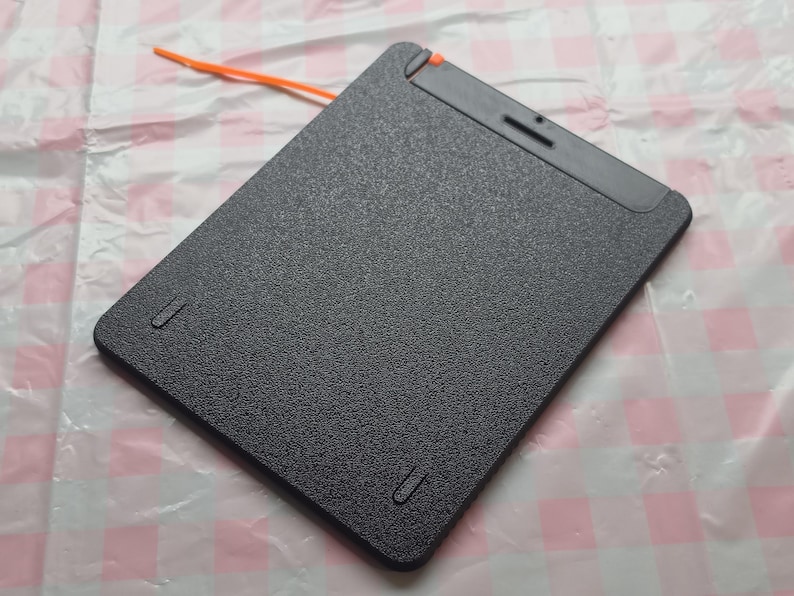 May include: A black, rectangular tablet with a textured surface. The tablet has a small orange cord attached to the top edge. There are two small, recessed slots on the bottom edge and one on the top edge.