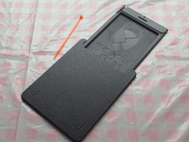 May include: A dark gray rectangular case with a sliding top, featuring the text "XIPH.COM ID: 8191". An orange stick lies beside the case on a pink and white checkered surface. The case appears to be made of plastic.