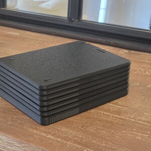 May include: A stack of six black, rectangular, textured plastic plates. Each plate has a slightly raised edge and a textured surface. The plates are stacked neatly on a wooden surface, with a dark-framed mirror in the background.