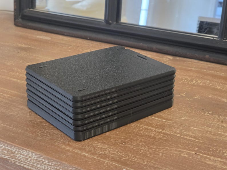 May include: A stack of six black, rectangular, textured plastic plates. Each plate has a slightly raised edge and a textured surface. The plates are stacked neatly on a wooden surface, with a dark-framed mirror in the background.