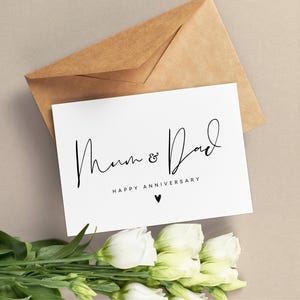May include: A white card with the words "Mum & Dad HAPPY ANNIVERSARY" in black script, with a small heart. The card is on a beige surface, with a brown envelope behind it and white flowers in the foreground.