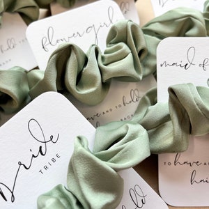 Peut inclure: Des chouchous vert sauge avec des cartes blanches qui disent "Bride Tribe", "Maid of Honor", "Flower Girl", et "To have and to hold hair back".
