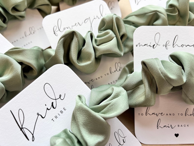 Peut inclure: Des chouchous vert sauge avec des cartes blanches qui disent "Bride Tribe", "Maid of Honor", "Flower Girl", et "To have and to hold hair back".