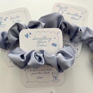 Puede incluir: Varios coleteros de sat&eacute;n azul claro con tarjetas blancas. Las tarjetas tienen ilustraciones azules y textos como "Bride to Be", "Something Blue" y "Something Blue Crew".