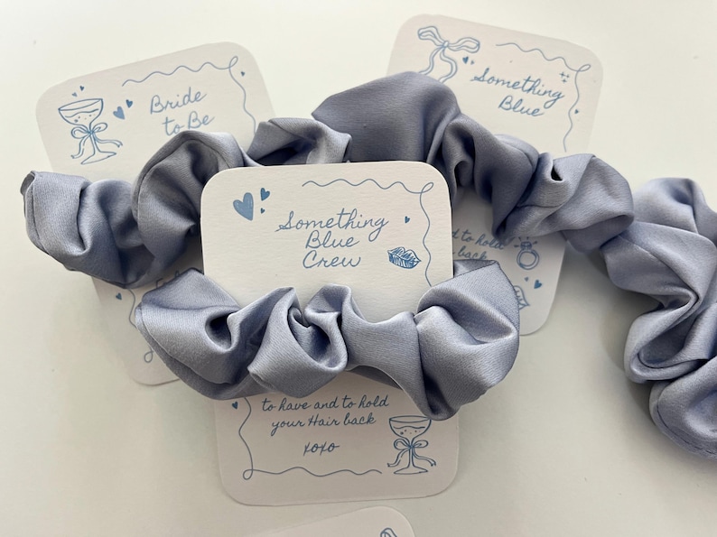 Puede incluir: Varios coleteros de sat&eacute;n azul claro con tarjetas blancas. Las tarjetas tienen ilustraciones azules y textos como "Bride to Be", "Something Blue" y "Something Blue Crew".